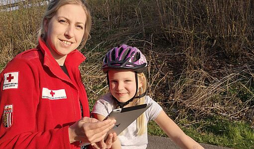 Jugendrotkreuz-Mitarbeiterin mit Tablet in der Hand neben einer Volksschülerin, die auf dem Rad sitzt