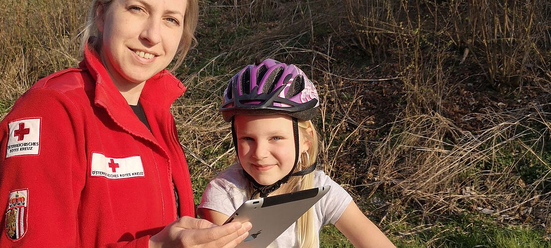 Jugendrotkreuz-Mitarbeiterin mit Tablet in der Hand neben einer Volksschülerin, die auf dem Rad sitzt