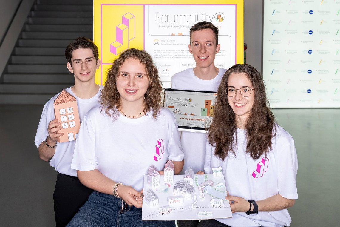 Vier Personen stehen vor einem Bildschirm mit „ScrumpliCity“ und halten ein gebasteltes Stadtmodell sowie ein Tablet in den Händen.