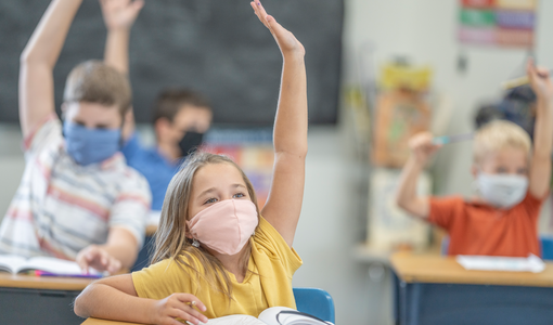 Volksschulkinder in der Klasse mit Maske, die gerade aufzeigen