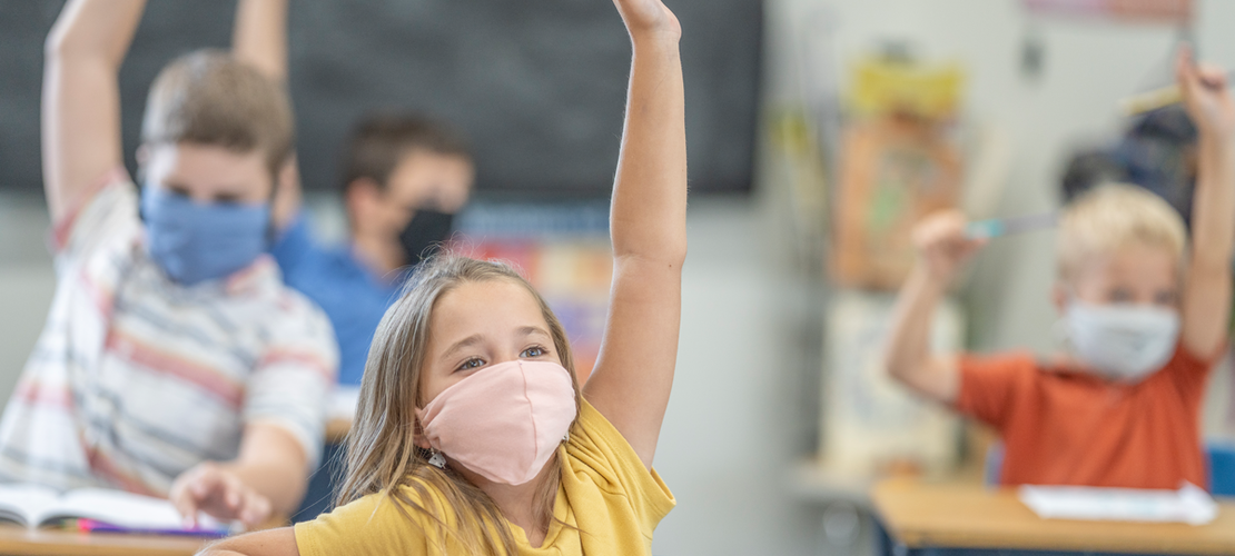 Volksschulkinder in der Klasse mit Maske, die gerade aufzeigen