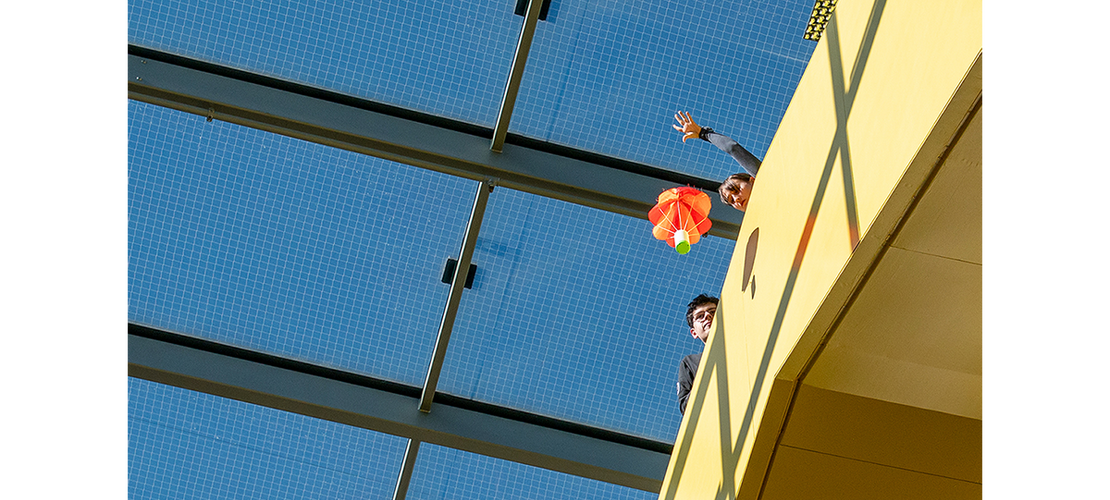 Von einer Balustrade unter einem gerasterten Glasdach lässt eine Hand einen kleinen Fallschirm mit einer grünen Kapsel in die Tiefe fallen, während zwei Jugendliche gespannt hinterherblicken.
