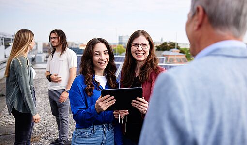 Zwei Schülerinnen sprechen mit einem Fachhochschulprofessor. Im Hintergrund spricht ein Schülerin mit einem Student.