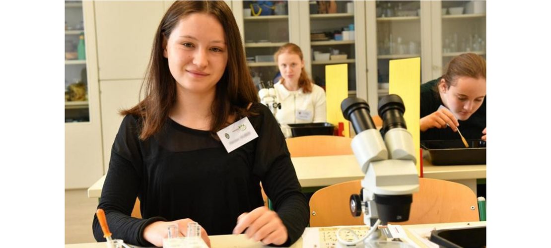 Schülerin bei einem Experiment im Biologiesaal, vor ihr ein Mikroskop; im Hintergrund arbeiten zwei weitere Schülerinnen.