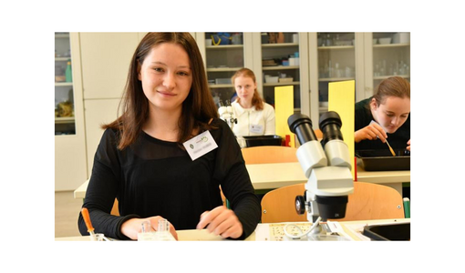 Schülerin bei einem Experiment im Biologiesaal, vor ihr ein Mikroskop; im Hintergrund arbeiten zwei weitere Schülerinnen.