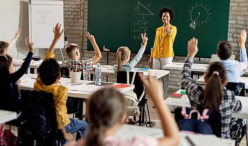 Kinder während des Unterrichts mit Lehrerin, einige zeigen auf