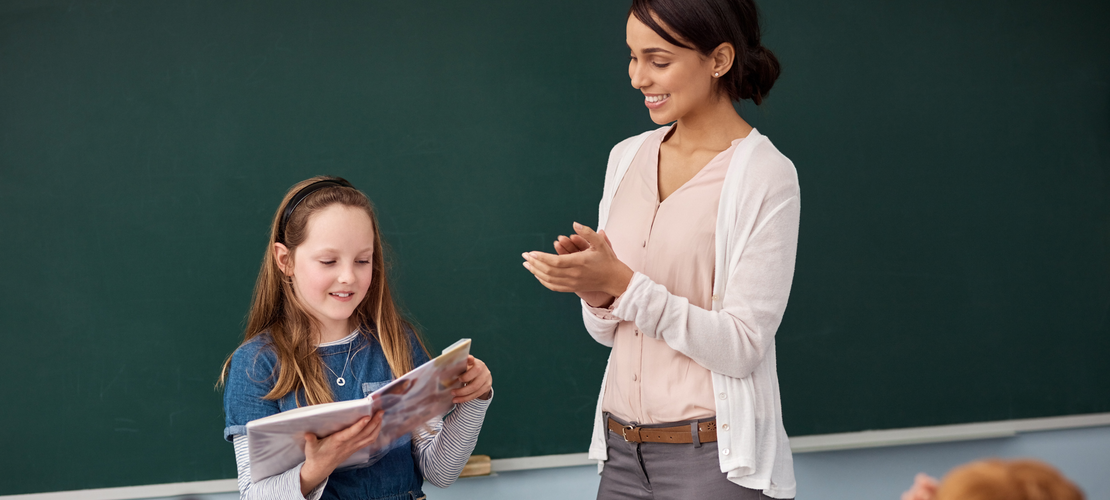 Schülerin steht vor der Tafel und trägt was vor, Lehrerin steht daneben und klatscht