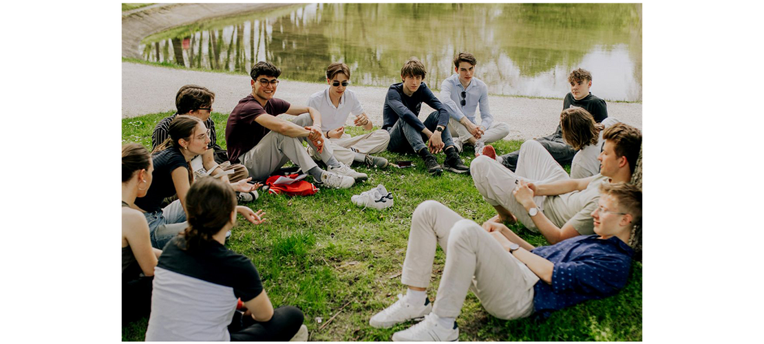Jugendliche sitzen im Kreis auf einer Wiese am Wasser und unterhalten sich.