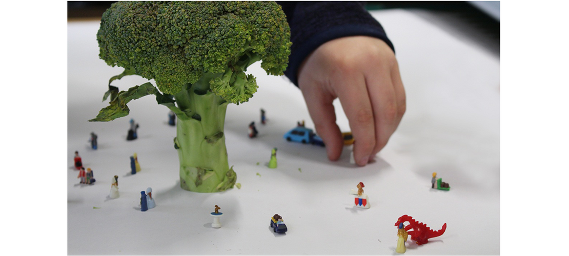 Eine Kinderhand stellt kleine Figuren und Fahrzeuge um einen großen Brokkoli auf einem weißen Tisch.