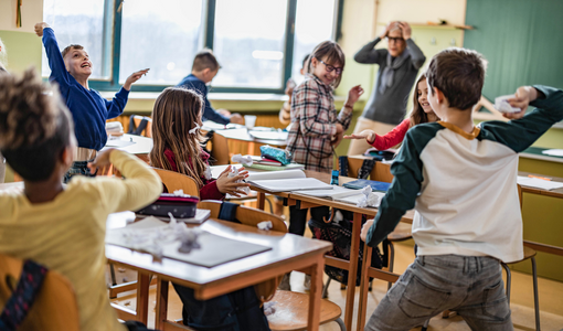 Eine große Gruppe von Schulkindern macht während eines Unterrichts im Klassenzimmer Chaos, während ihr Lehrer darüber frustriert ist.