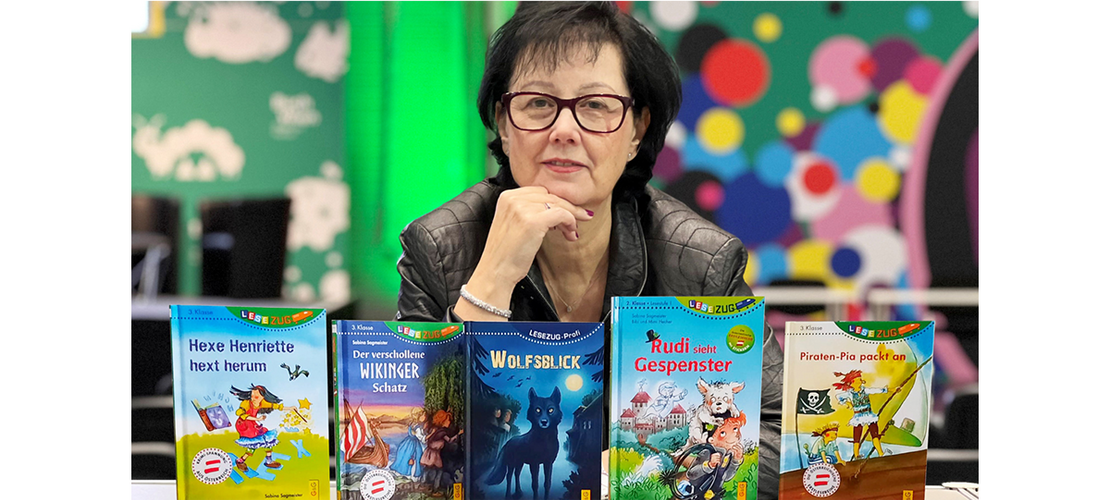 Sabina Sagmeister sitzt hinter mehreren Kinderbüchern und blickt in die Kamera; bunte Bücher stehen auf dem Tisch vor ihr.