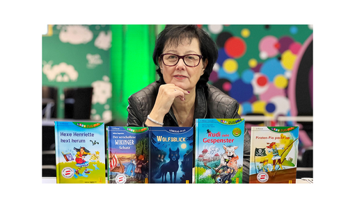 Sabina Sagmeister sitzt hinter mehreren Kinderbüchern und blickt in die Kamera; bunte Bücher stehen auf dem Tisch vor ihr.